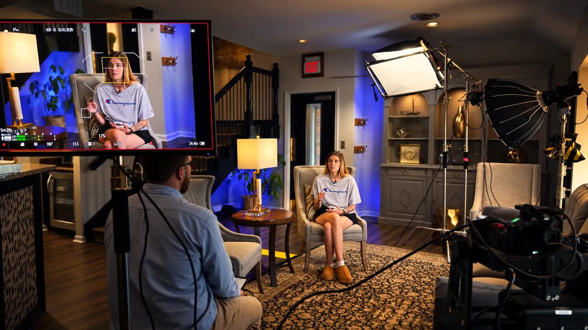 An interview set styled as a living room: a camera monitor frames a young woman in a grey T-shirt seated in an armchair, while an interviewer faces her under softbox and parabolic lights.